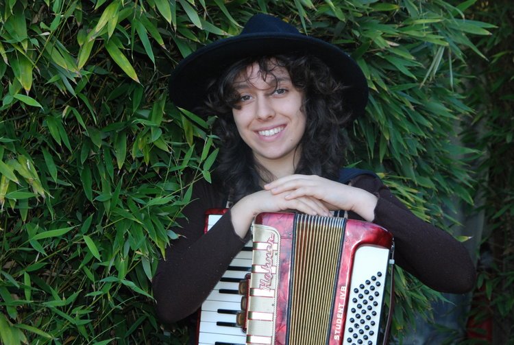   Leavy smiles brightly against a backdrop of flourishing greenery, her hands comfortably crossed atop her accordion.   