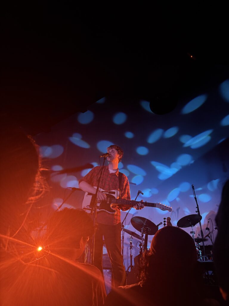 Image shows Dove Ellis playing guitar center stage in front of a drum set, a few people can be seen.